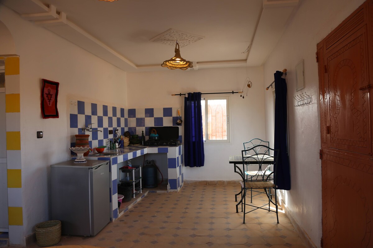 The kitchen area features vibrant blue and white tiles, complemented by wooden cabinetry. A compact round table with two metal chairs is situated near a window framed by dark curtains. Various kitchen appliances, including a refrigerator, are visible, providing functional space for meal preparation.