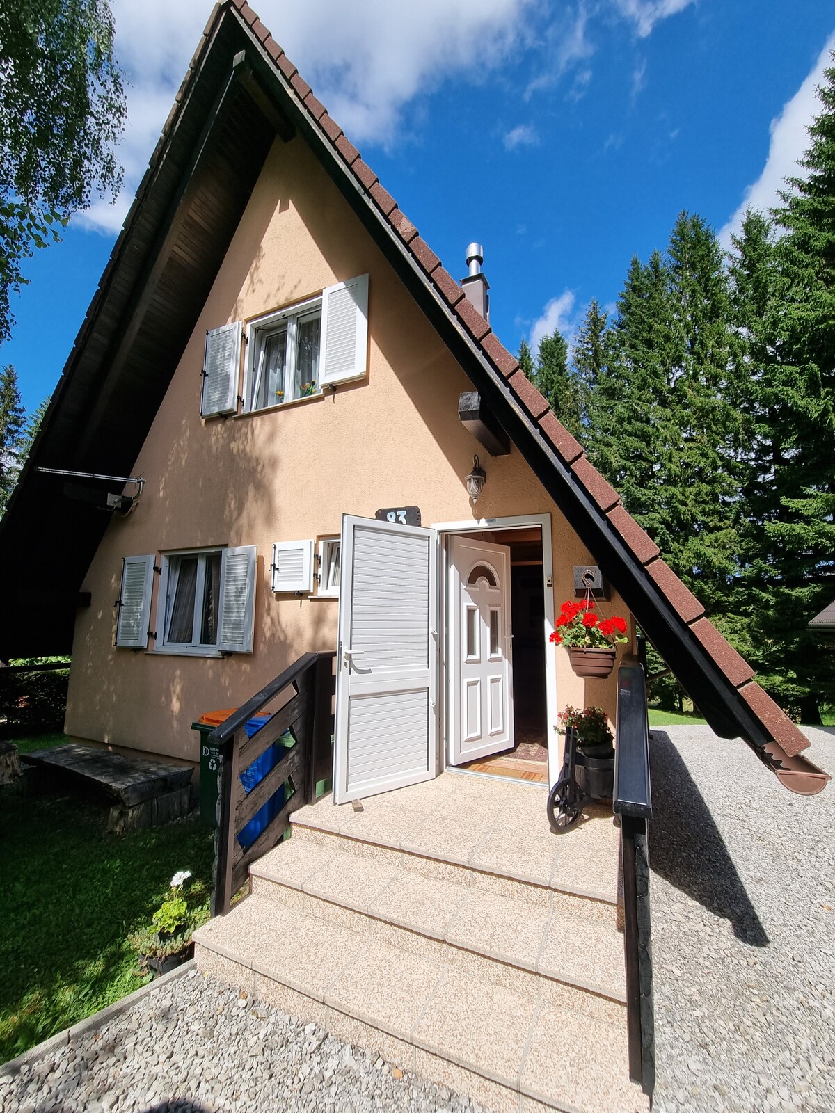 The charming mountain hut features a distinctive triangular roof and a stone walkway leading to the entrance. Bright flower arrangements adorn the front, while surrounding greenery enhances the peaceful setting. Windows with shutters complement the warm exterior, inviting guests to explore the serene surroundings.
