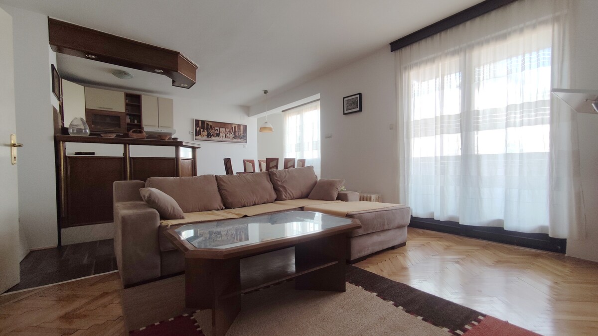 A spacious living area features a light-colored sectional sofa arranged around a low wooden coffee table. Large windows allow natural light to flood the room, with sheer curtains softly diffusing the sunlight. The open-concept space includes a view of the kitchen area in the background.