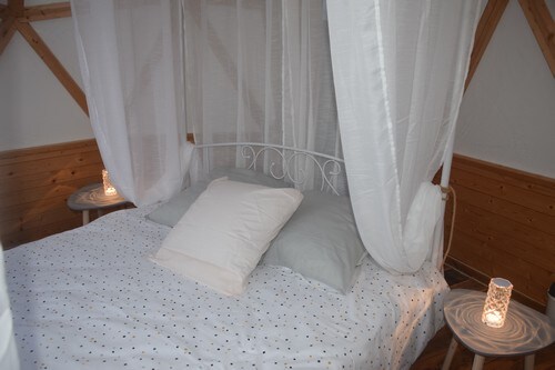 A cozy sleeping area features a white metal bed with a light-colored bedspread adorned with small dots. Soft pillows are arranged on the bed, and warm light emits from two side tables with decorative candles, creating a serene and intimate atmosphere.