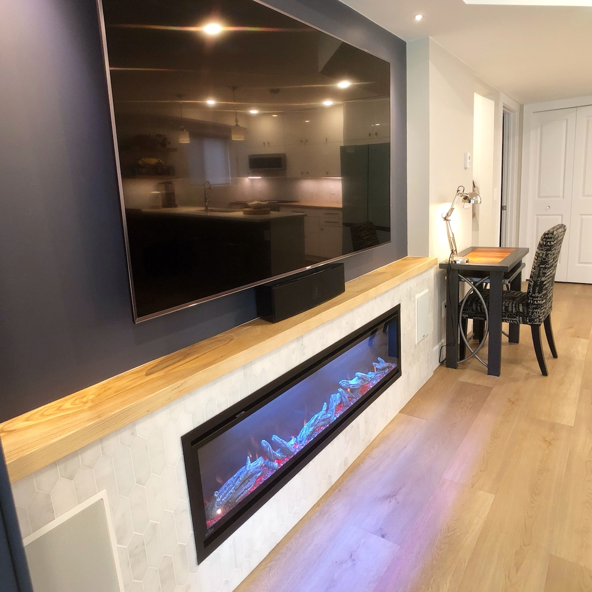 An open living area features a large wall-mounted television above an electric fireplace, which displays colorful lights. A light wood shelf runs beneath the television, complemented by a small desk with a chair and lamp positioned nearby, enhancing the functional space.