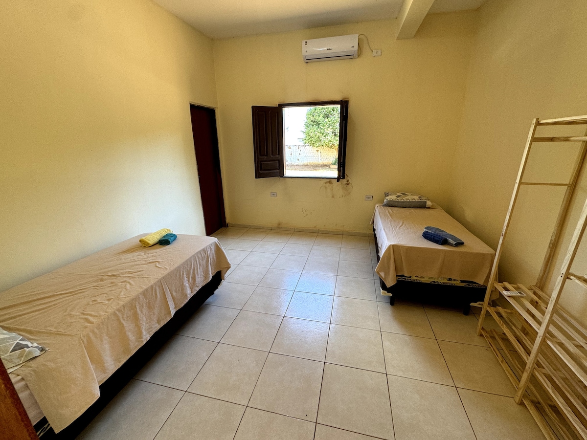 A bright room features two twin beds with light-colored linens. An air conditioning unit is mounted on the wall, providing comfort. Natural light enters through a window, which is adorned with wooden shutters. A bamboo rack stands against the wall, offering additional storage space.