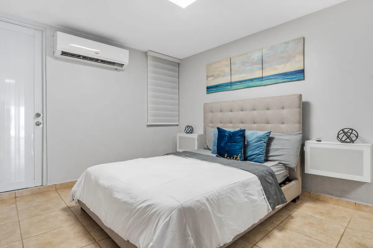 A cozy bedroom features a spacious bed adorned with light bedding and decorative pillows in blue tones. Artwork depicting a serene landscape hangs above the headboard. Soft lighting from an overhead fixture enhances the calm ambiance, while large windows are fitted with light-filtering shades.