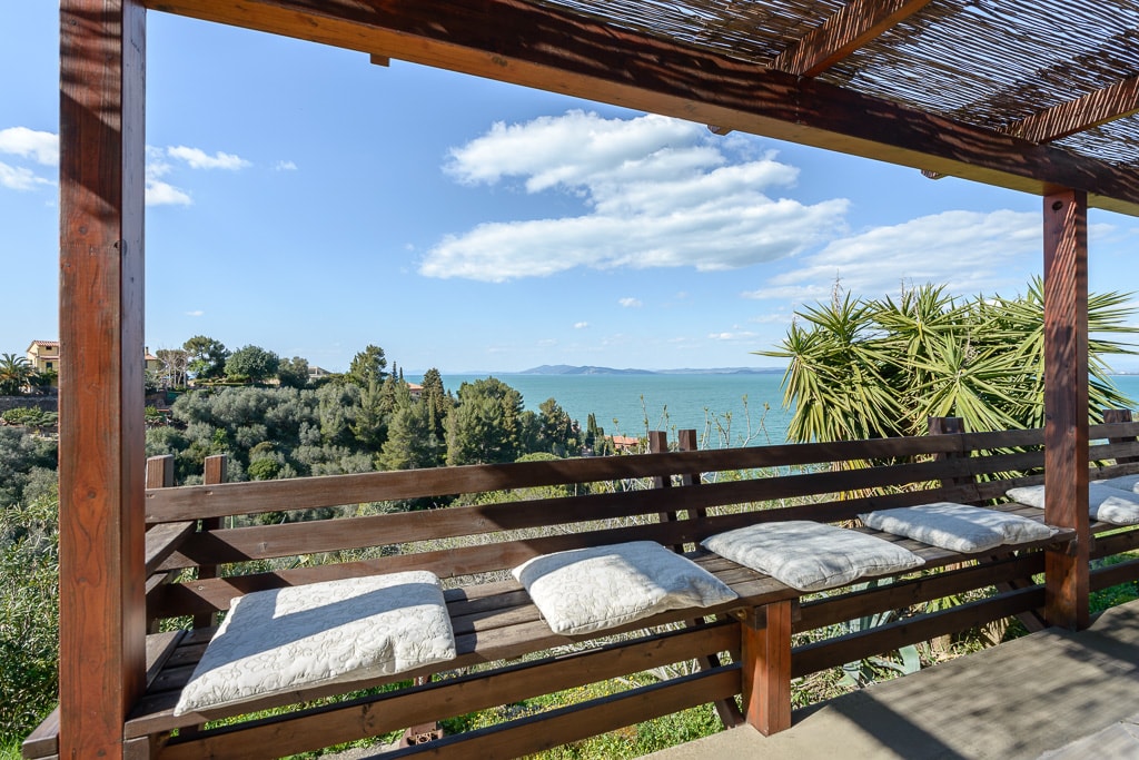 A covered outdoor seating area features a long wooden bench adorned with soft cushions. The space offers a clear view of the sea and nearby greenery, allowing for a peaceful atmosphere. The surrounding natural landscape complements the inviting seating arrangement.