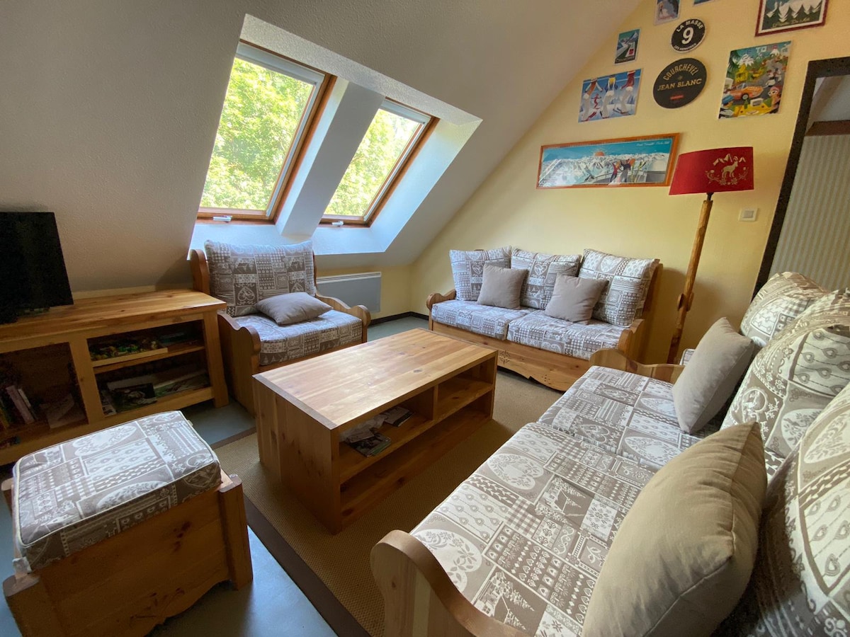A comfortable sitting area is presented with two wooden sofas adorned with patterned cushions. A low wooden coffee table sits in the center, complemented by a matching ottoman. Slanted windows allow natural light to fill the space, enhancing the inviting decor on the walls.