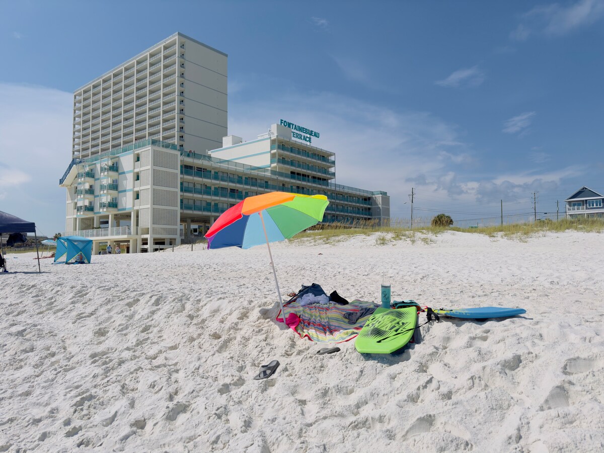 A vibrant beach scene features colorful umbrellas along the white sand. The background showcases the tall, modern Fontainebleau Terrace building, providing a clear view of beachfront access. Surfboards and towels are neatly arranged near the vibrant umbrella, inviting relaxation by the water.