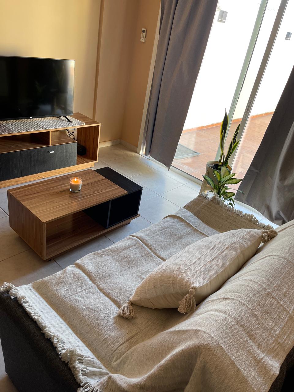 A comfortable living area is presented, featuring a dark sofa adorned with a light throw blanket. A wooden coffee table is placed in front, complemented by a small plant and a candle. Natural light enters through glass doors leading to an outdoor space.