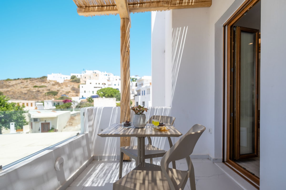 An outdoor terrace features a wooden table accompanied by light-colored chairs, inviting relaxation. A potted plant adds a touch of greenery. Sunlight filters through a thatched overhang, creating a bright atmosphere while offering scenic views of nearby buildings and hills.