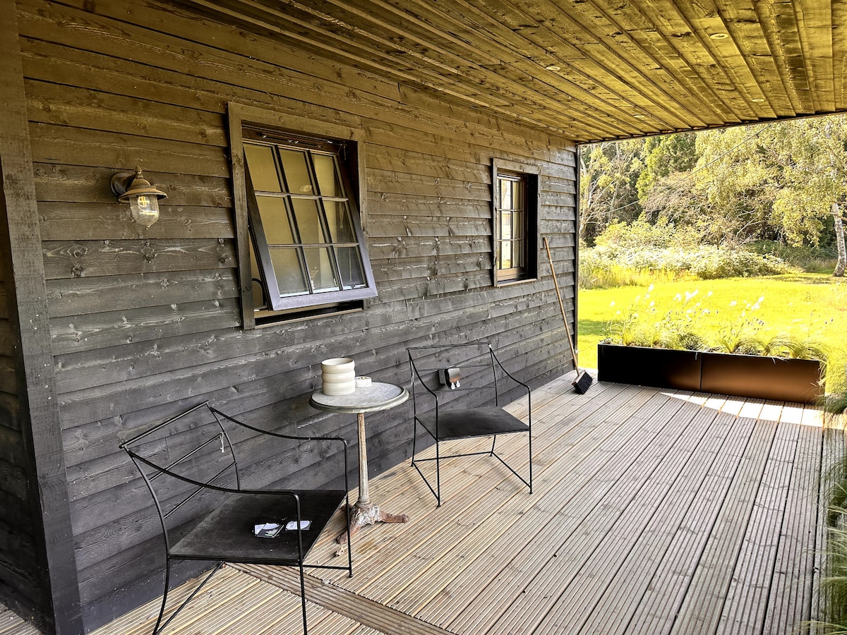 An outdoor deck area features a wooden floor and a small circular table paired with two black wire chairs. Natural light streams through the window, and lush greenery is visible beyond the railing, highlighting the serene outdoor setting.