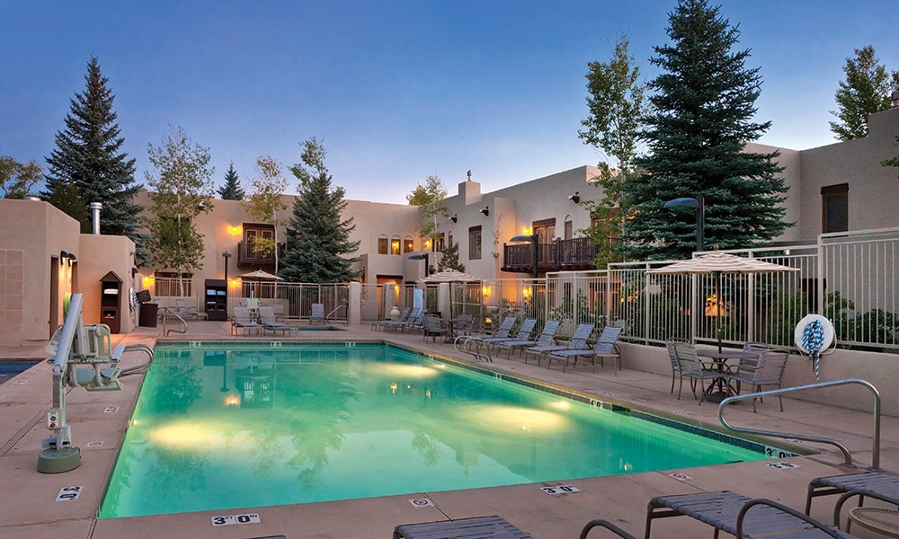 An outdoor swimming pool glows under the evening sky, surrounded by lounge chairs and shaded by tall trees. The resort buildings, styled in a pueblo design, are visible in the background, reflecting a warm atmosphere with soft lighting.