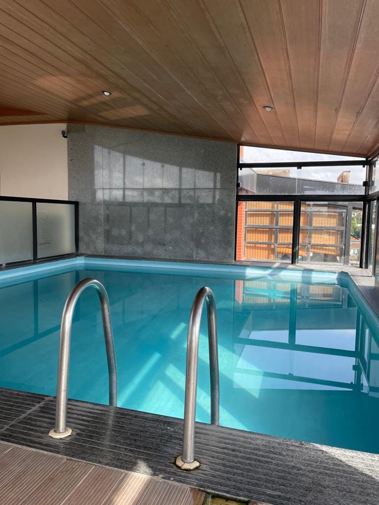 An indoor pool is featured within a well-lit space, showcasing clear turquoise water. Natural light filters through large windows, reflecting off the smooth granite walls and wooden ceiling. Stainless steel handrails are positioned at the pool's edge for safety.