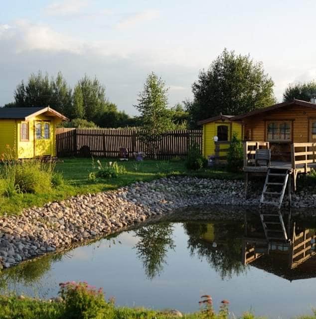 Two yellow wooden cabins are situated beside a serene pond, reflecting their vibrant color. Lush greenery surrounds the area, with trees and plants offering a natural setting. A wooden deck leads to one cabin, enhancing the peaceful, rural atmosphere.
