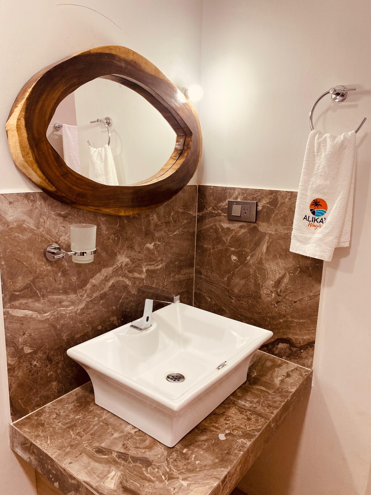 A modern bathroom sink is displayed, featuring a square white basin atop a brown marble countertop. A circular wooden mirror adds warmth to the space, while a towel hangs neatly on a chrome holder nearby.