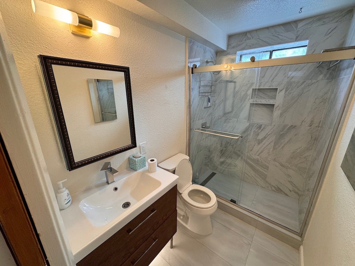 A modern bathroom is presented with marble-patterned walls and a glass-enclosed standing shower. A sleek vanity with a single sink and dark wood cabinetry is complemented by a large mirror. Natural light enters from a nearby window, enhancing the clean and spacious feel of the area.