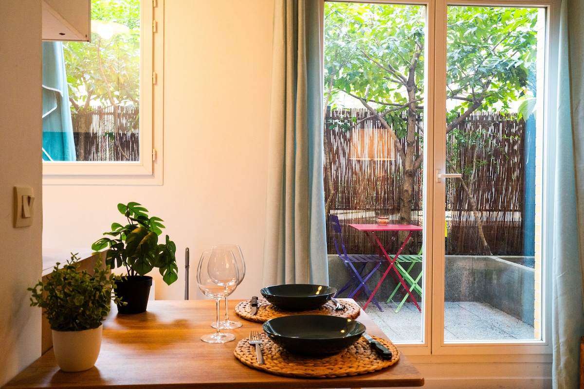 A dining area features a wooden table set with two green plates and wine glasses. Sunlight filters through sheer curtains, illuminating a potted plant. The view through glass doors reveals a private terrace with colorful outdoor seating.