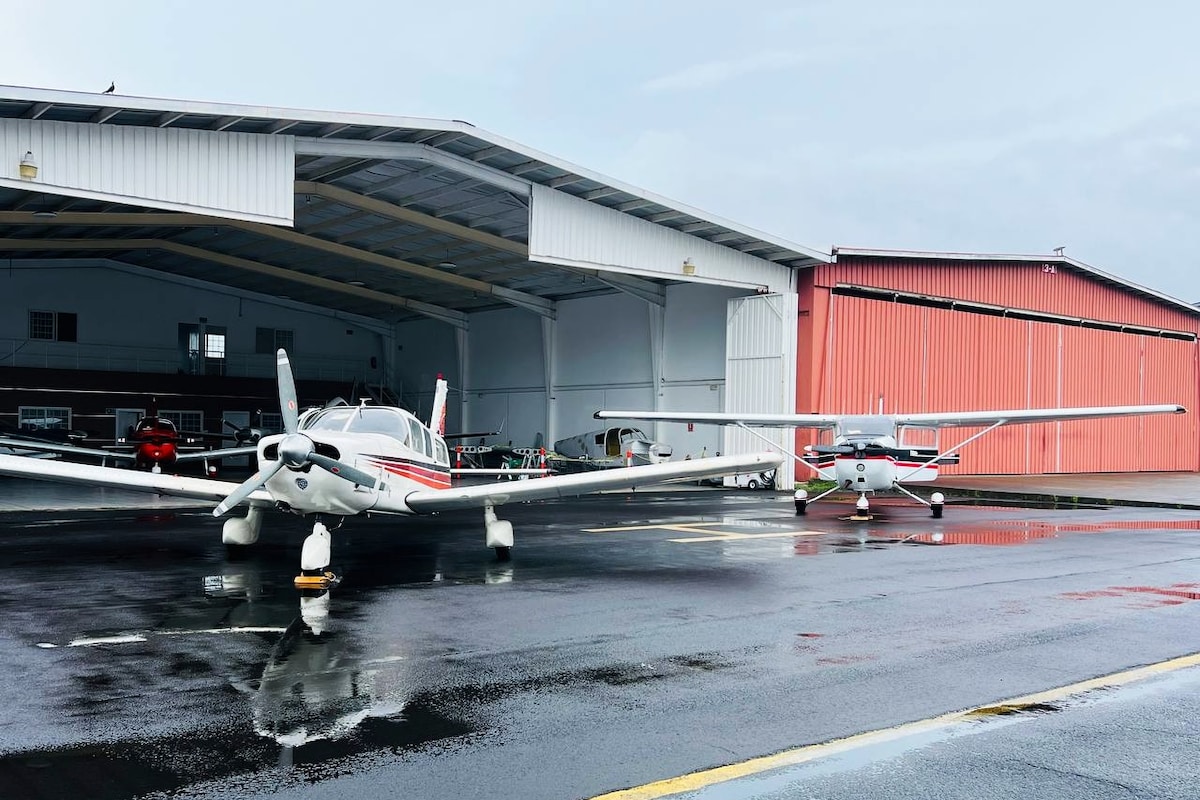 Two aircraft are positioned on a wet runway in front of a hangar. A larger plane with red and white detailing is situated prominently, while a smaller plane is parked nearby. The hangar structures are visible in the background, showcasing a mix of metal and wood designs.