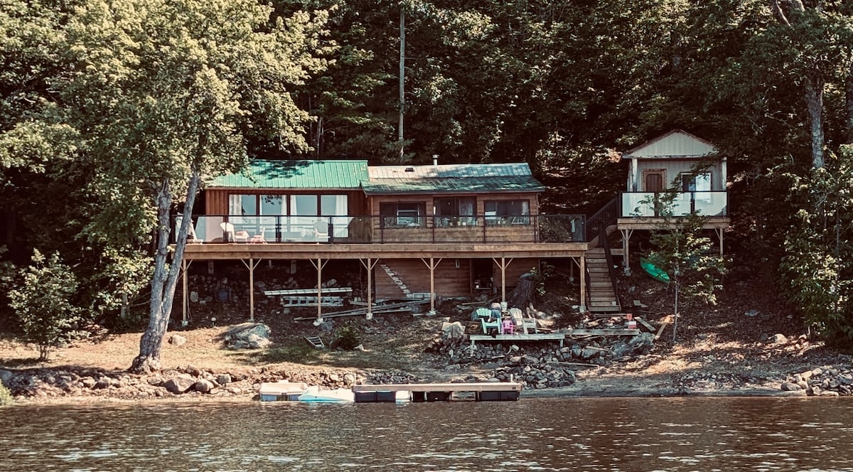 The rustic cabin, with a green metal roof, is set among lush trees by the water's edge. A spacious deck runs along the front, providing access to seating areas. A separate bunkie is visible to the right, connected by a short set of stairs.