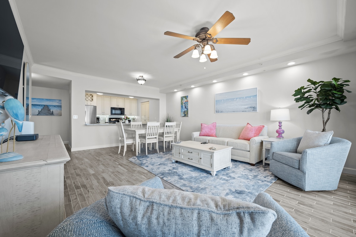 A bright living space features a light-colored sofa and armchair, adorned with pink and blue cushions. A wooden coffee table sits centered on a patterned blue area rug. The open layout includes a dining area with white chairs, and the kitchen is visible in the background.