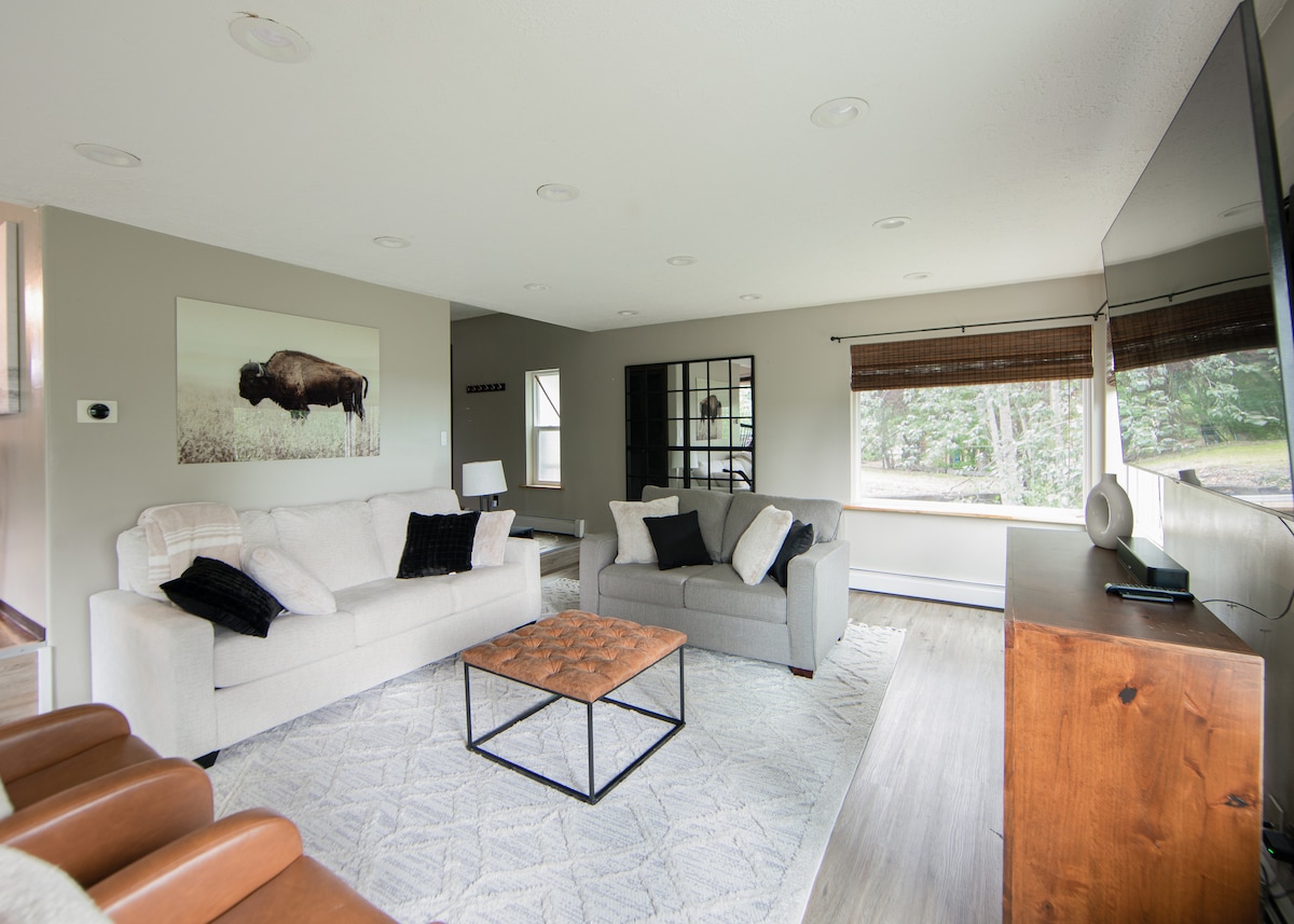 A spacious living room features a light sofa and a gray sectional, complemented by a coffee table with a geometric design. Large windows provide natural light and views of the outside greenery. A wall-mounted television and decorative artwork enhance the room's modern aesthetic.