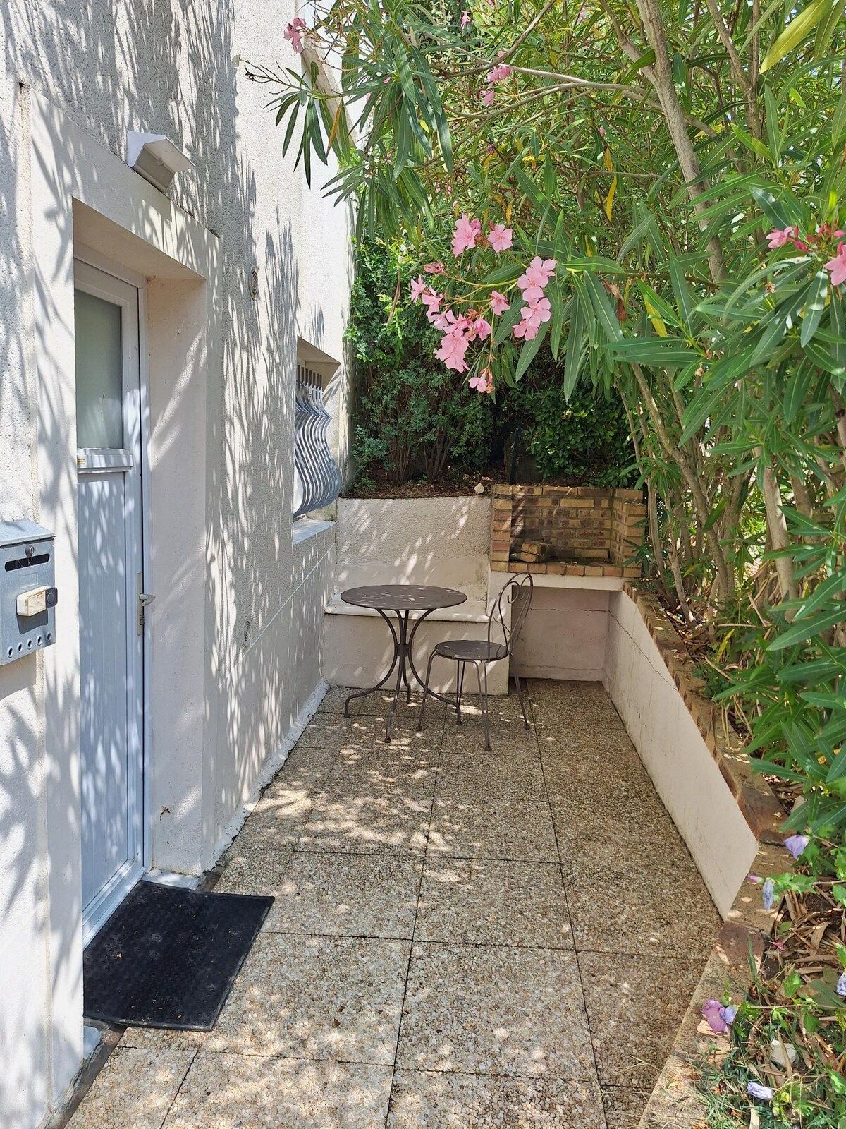 A small private terrace is visible adjacent to the studio entrance, featuring a round table and two chairs. Lush vegetation, including flowering plants, surrounds the area, providing a sense of privacy and nature. The gravel surface is well-maintained, enhancing outdoor relaxation.