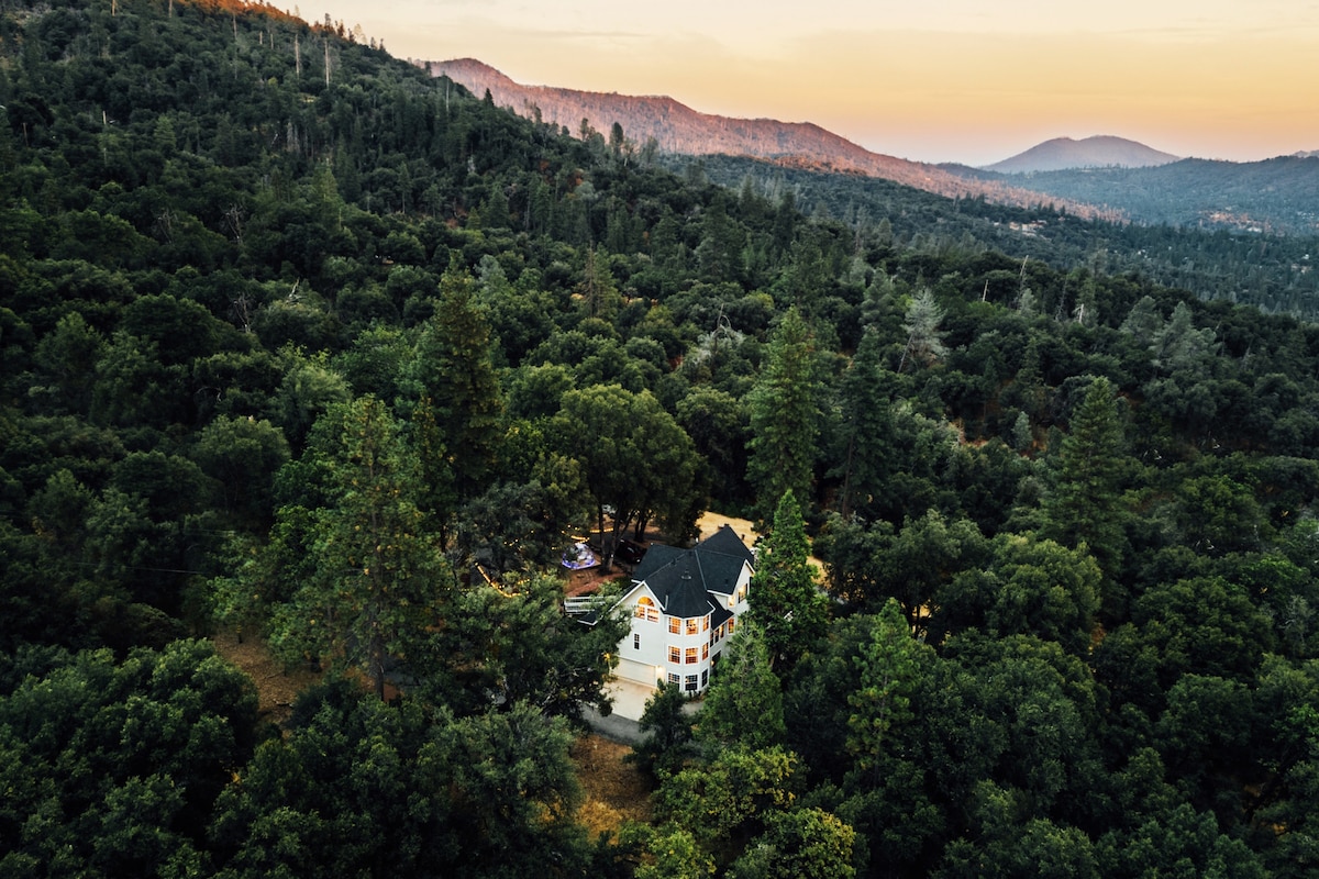The spacious retreat is set against a backdrop of lush greenery and rolling mountains. The aerial view captures the surrounding trees and the home’s inviting exterior, showcasing a blend of natural beauty and architectural charm. The distant horizon reveals a soft sunset glow.