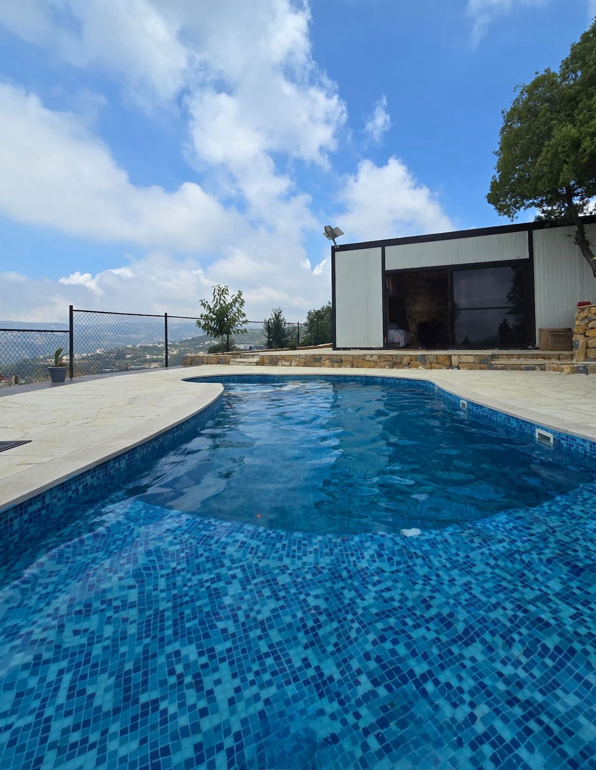 A private pool is shown, featuring a clear blue mosaic design. Surrounding the pool is a natural stone patio, with a safety fence in the background. A modern container structure is visible nearby, complemented by greenery and an expansive sky with fluffy clouds.