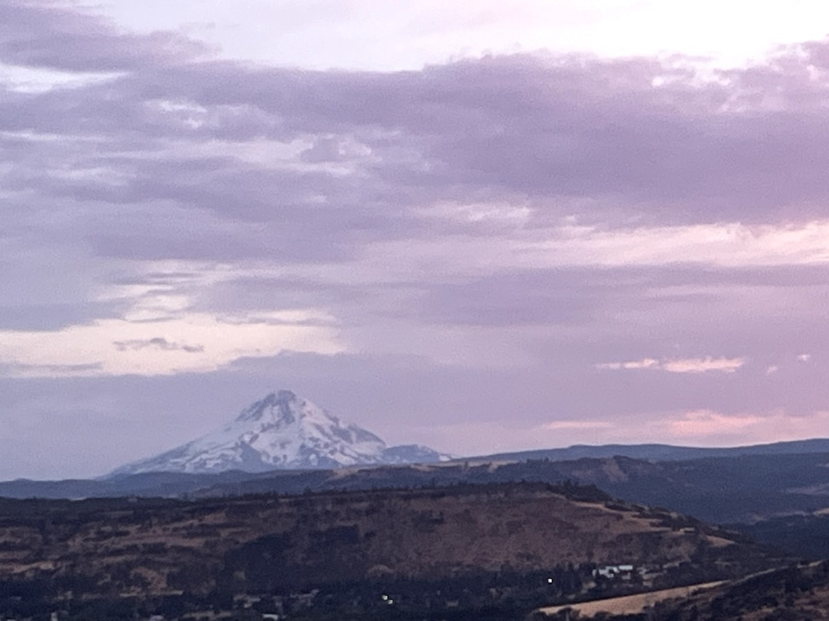 A serene view of Mt. Hood is captured under a pastel sky, with clouds reflecting shades of purple and pink. The mountain rises majestically above the surrounding landscape, revealing patches of snow on its peak against a backdrop of rolling hills.