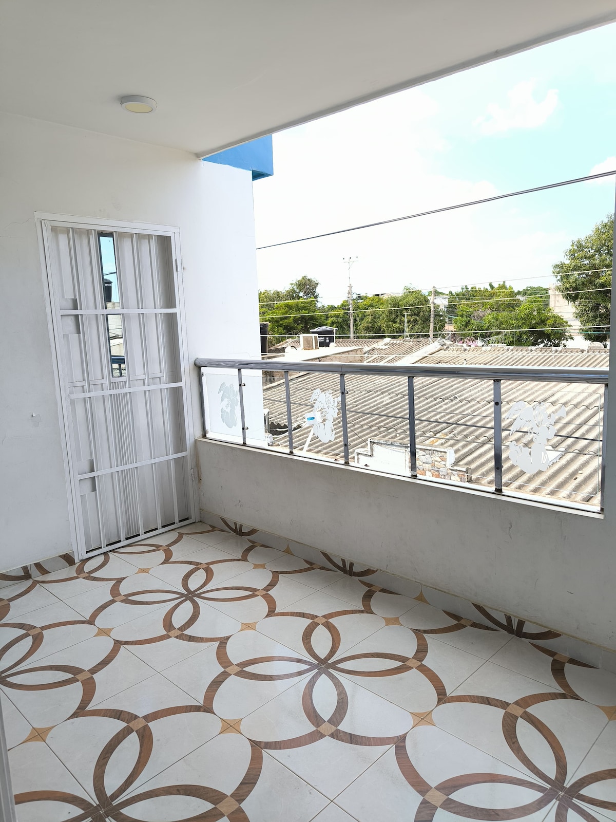 A balcony space with patterned tile flooring is visible, featuring a glass railing that offers a view of the outside. Natural light fills the area, framed by a white door and a nearby window. Surrounding greenery and rooftops can be seen in the background.