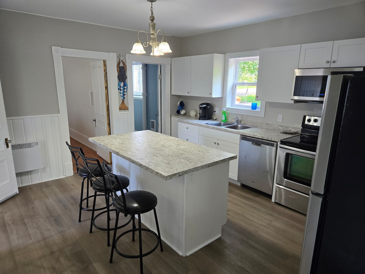 A modern kitchen features white cabinetry and stainless steel appliances. A multi-functional island with seating for three is positioned at the center. Natural light enters through the window above the sink, enhancing the light grey walls and wooden flooring.