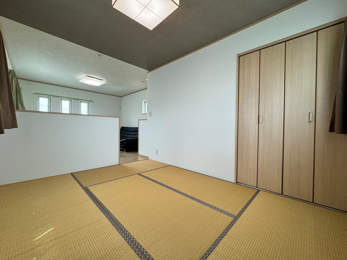 A clean and spacious room features traditional tatami flooring, providing a sense of serenity. A storage closet is present on one side, while a window allows natural light to fill the space. An inviting seating area is visible in the background, enhancing its functionality.