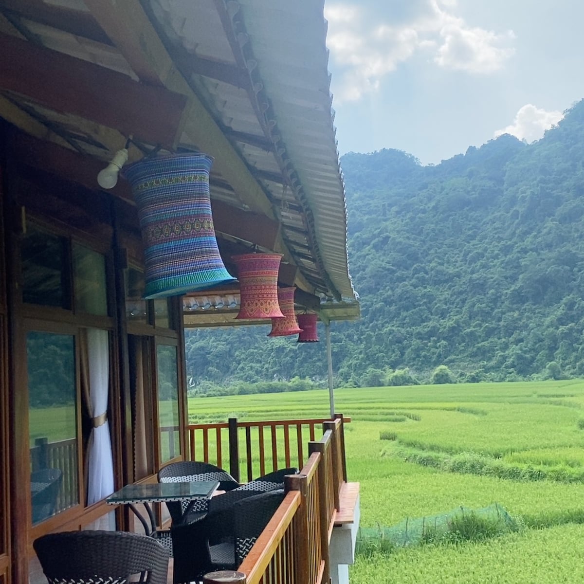 A balcony features several colorful woven lanterns hanging from the roof, overlooking lush green rice fields and distant rolling hills. Black outdoor furniture is positioned on the balcony, offering a serene view of the surrounding landscape.