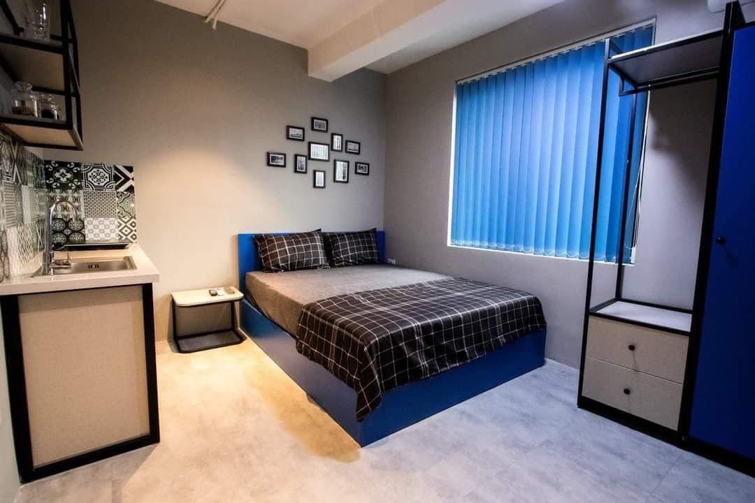 The studio apartment features a spacious bed with a patterned blanket, against a backdrop of light blue walls. A small kitchenette is presented in one corner, complemented by a geometric backsplash. Framed photographs adorn the wall, adding a personal touch to the minimalist design.