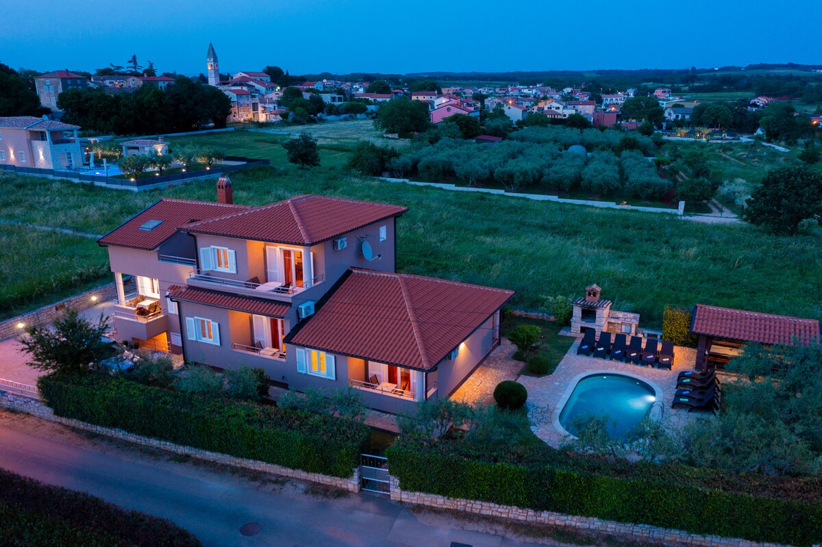 An aerial view of a house with a warm terracotta roof showcases a swimming pool surrounded by lounge chairs. The spacious property is set against lush greenery and a sunset sky, with neighboring buildings and rolling hills in the background.