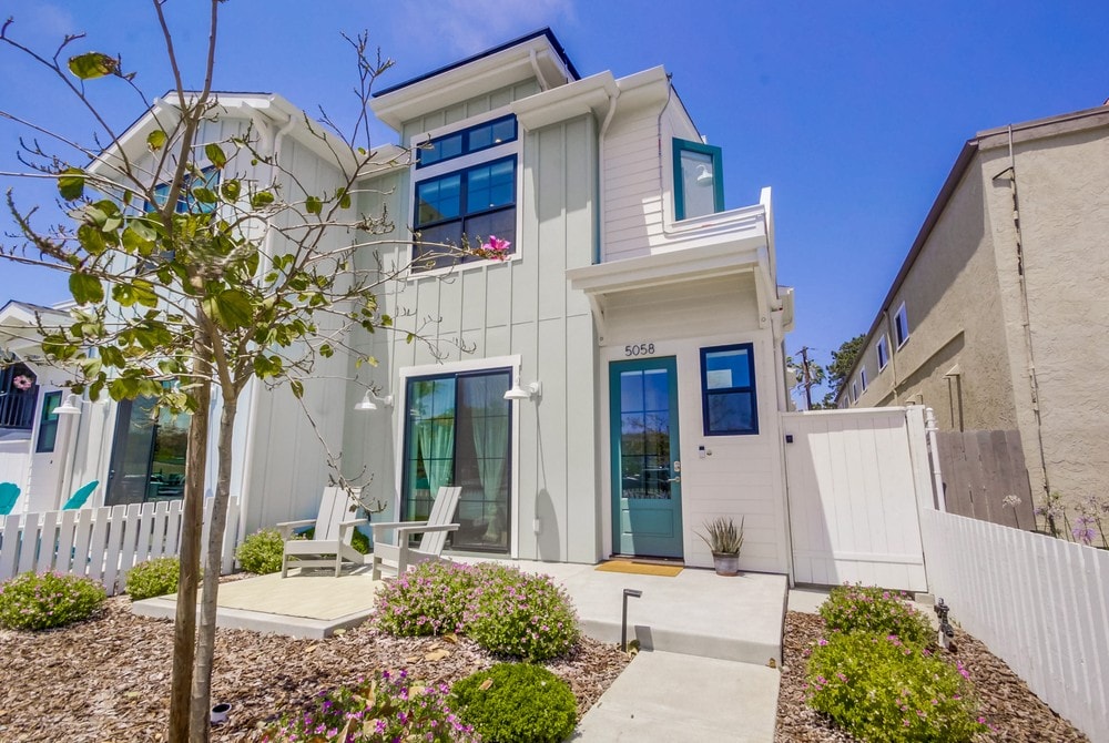 A modern, two-story beach house is displayed, featuring a light-colored exterior and a welcoming front porch. Two white chairs sit on a small patio, surrounded by landscaped gardens with green shrubs and flowering plants. The blue front door adds a vibrant touch.