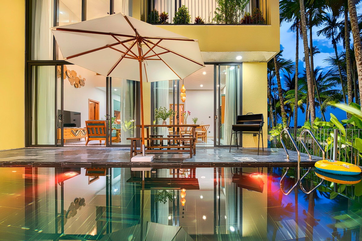 An outdoor seating area is visible beside a private pool, with a large umbrella providing shade. The calm water reflects the surrounding trees and open space. Glass doors lead to an interior area where dining and relaxation can be enjoyed, enhancing the connection between indoors and outdoors.