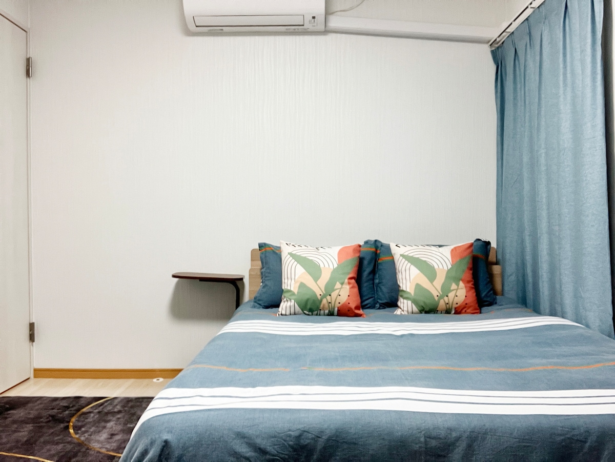 The sleeping area features a spacious bed dressed in a blue coverlet, complemented by decorative pillows in warm tones and tropical patterns. A small table is positioned next to the bed, while light blue curtains cover one window, enhancing the calm atmosphere of the room.