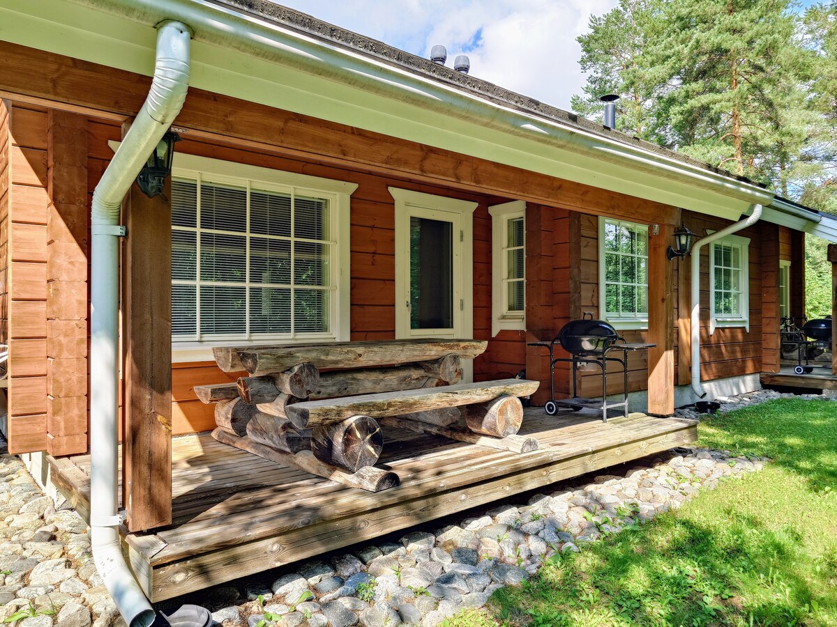 A wooden deck area features a large, rustic log dining table with attached benches, surrounded by greenery. A barbecue grill is positioned nearby, and oversized windows allow natural light to fill the space, complementing the warm wooden exterior.