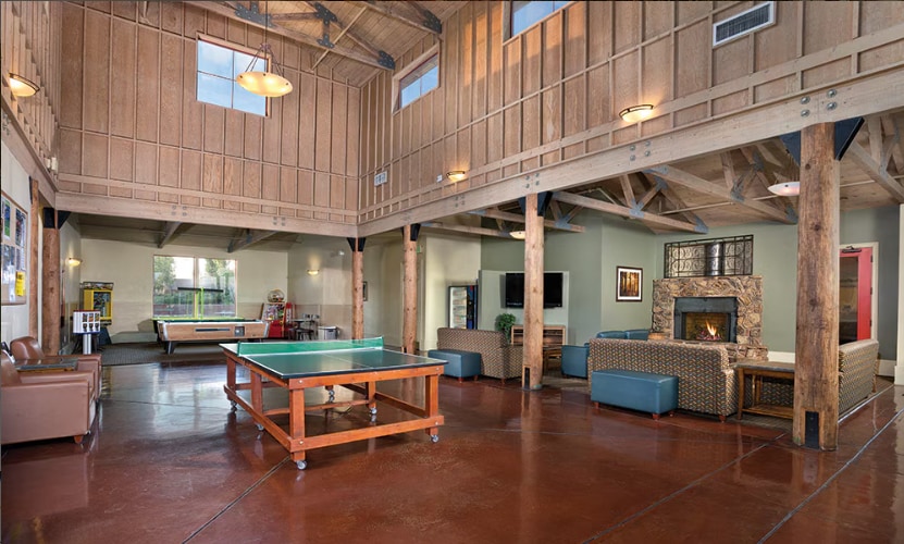 A spacious common area features high wood-beamed ceilings and large windows allowing natural light. A pool table is positioned centrally near seating areas with comfortable couches. A fireplace adds warmth to the space, complemented by a multimedia setup against the wall.