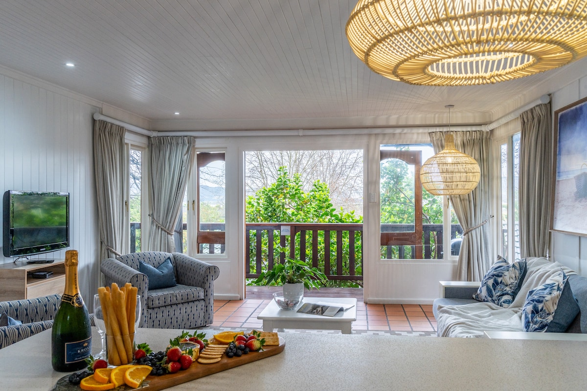 The living area features a cozy arrangement of seating including a patterned armchair and a light-colored sofa. A natural light source enters through large windows, framed by soft curtains. A festive snack board, complete with fruits and snacks, is positioned on the table. A television is mounted on the wall.