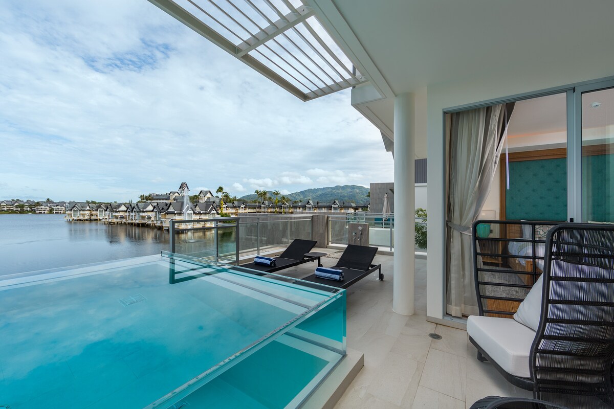 The private terrace features sun loungers overlooking a serene lake, complemented by a pool that mirrors the sky. The edge of the terrace is lined with glass barriers, providing unobstructed views of the tranquil water and surrounding greenery.