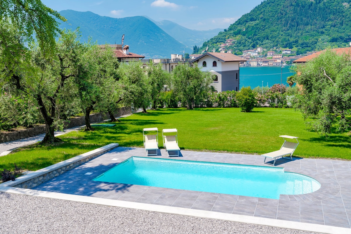 A private pool is surrounded by well-maintained grass and olive trees, offering a serene view of the mountains and lake. Lounge chairs are positioned beside the pool, providing a calm space for relaxation.