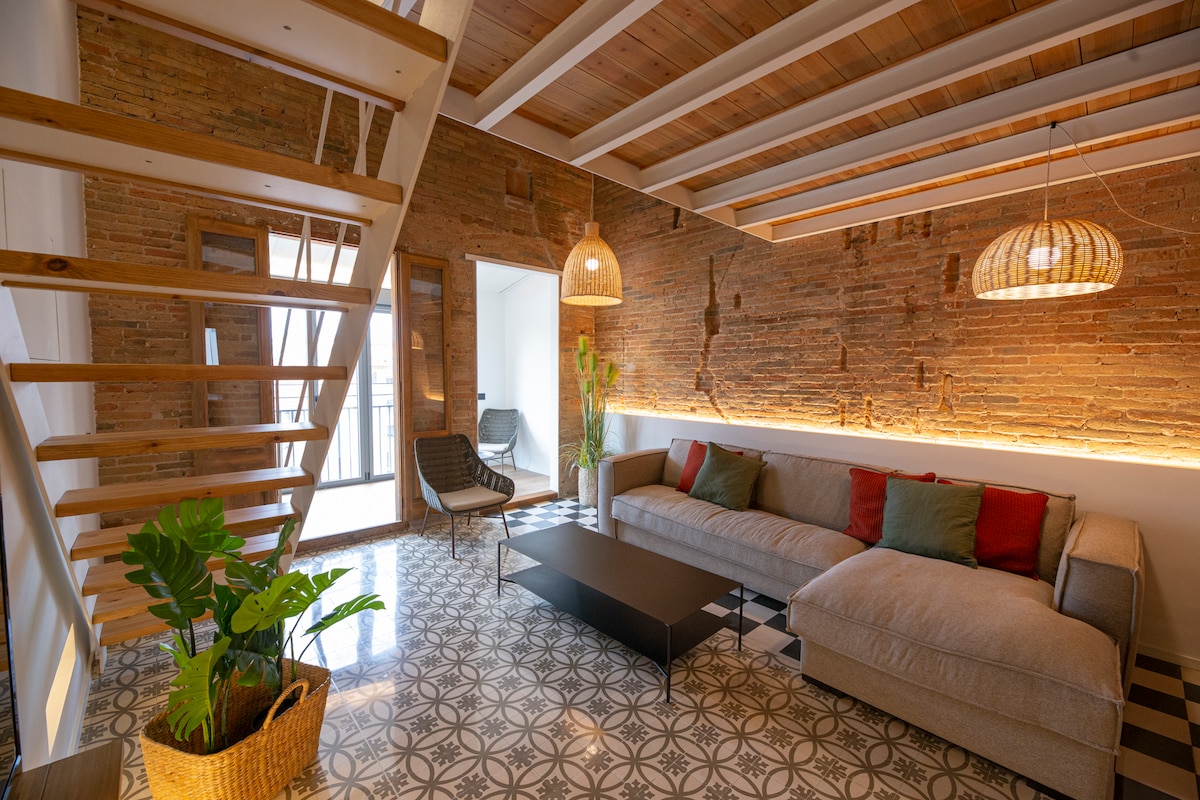 A spacious living area features a light gray sectional sofa with colorful accent pillows. The exposed brick wall contrasts with the patterned floor tiles. A staircase leads to an upper level, while pendant lights add a soft glow to the room.