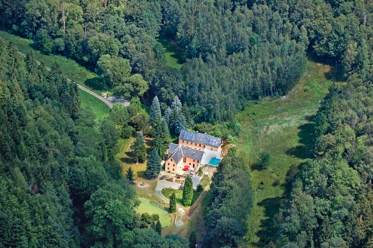 An aerial view captures the secluded Obere Trebnitzmühle set amidst lush greenery. The building is surrounded by dense forests, with a pool visible on the property, blending harmoniously with the natural landscape.