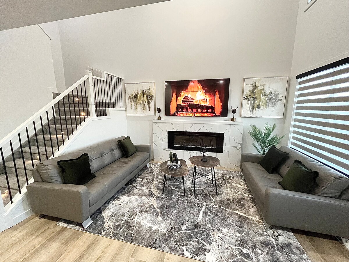 A modern living room is displayed, featuring two gray sofas placed on a plush area rug. A sleek coffee table sits between them. Above the fireplace, a large flat-screen TV is mounted, showcasing a warm, glowing fire. Art pieces adorn the walls, enhancing the contemporary design.