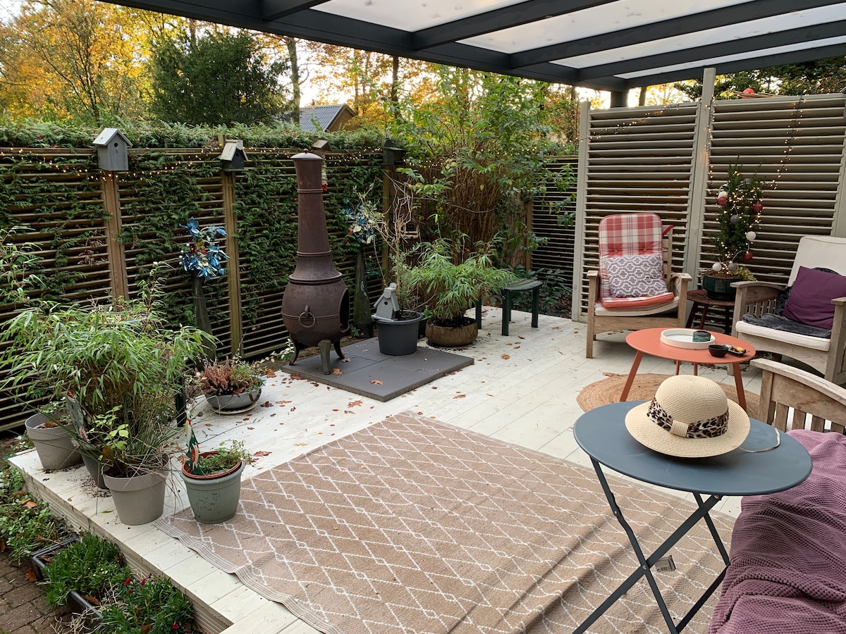 A covered outdoor deck is presented with a cozy seating arrangement, featuring a mix of wooden chairs and a small table. A patterned rug and decorative plants enhance the space, while a chiminea provides warmth. Surrounding greenery offers privacy in this tranquil area.