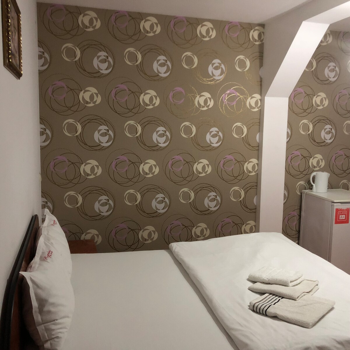 The sleeping area features a spacious bed with a simple headboard, accompanied by neatly arranged towels on the bed. A patterned wallpaper in soft tones provides visual interest, while a small appliance is seen in the corner, indicating additional accommodation features.