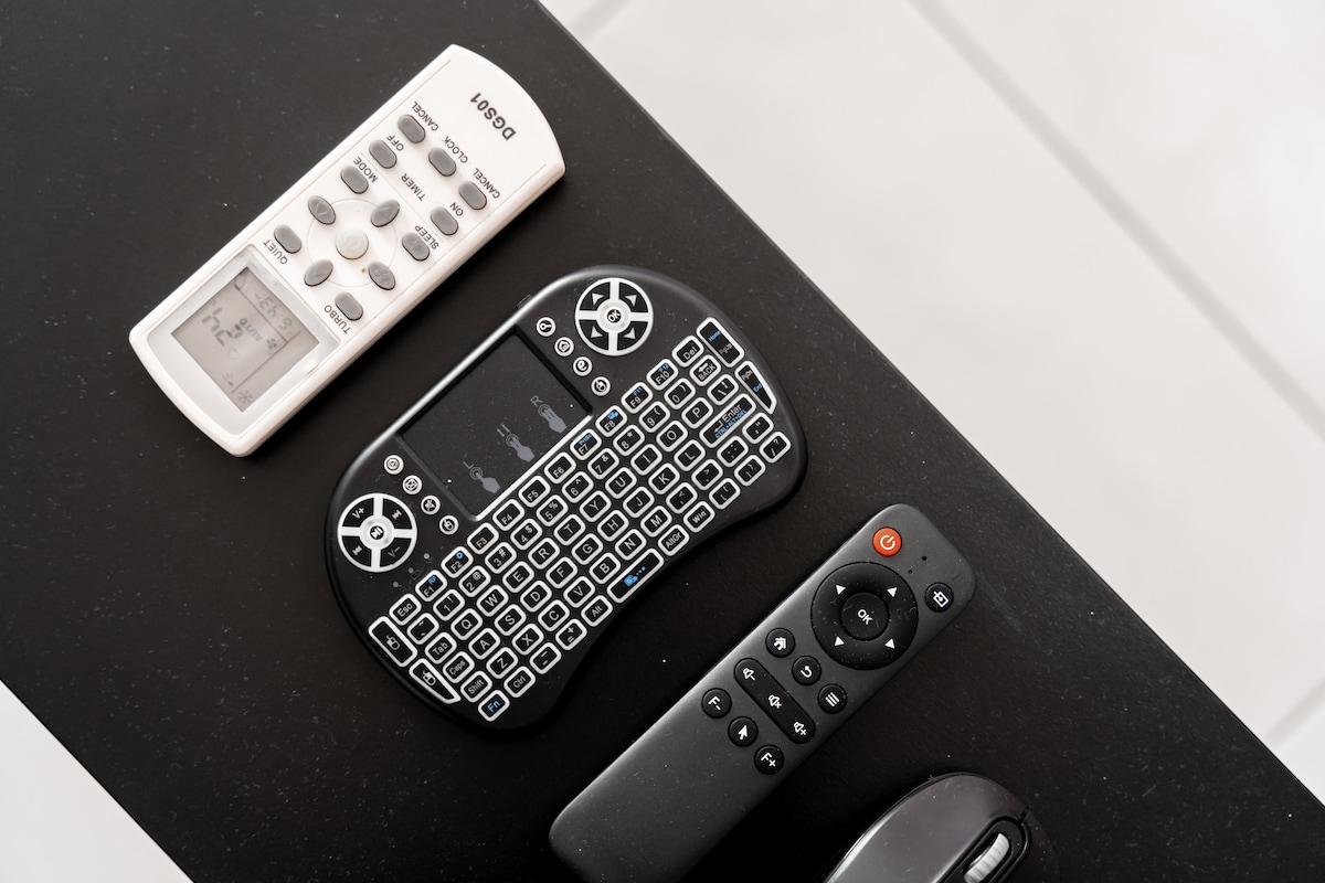 Three remote controls are neatly arranged on a black tabletop. They include a white remote with multiple buttons, a black wireless keyboard with a touchpad, and a sleek black remote with a power button. The surface is smooth and minimalist.