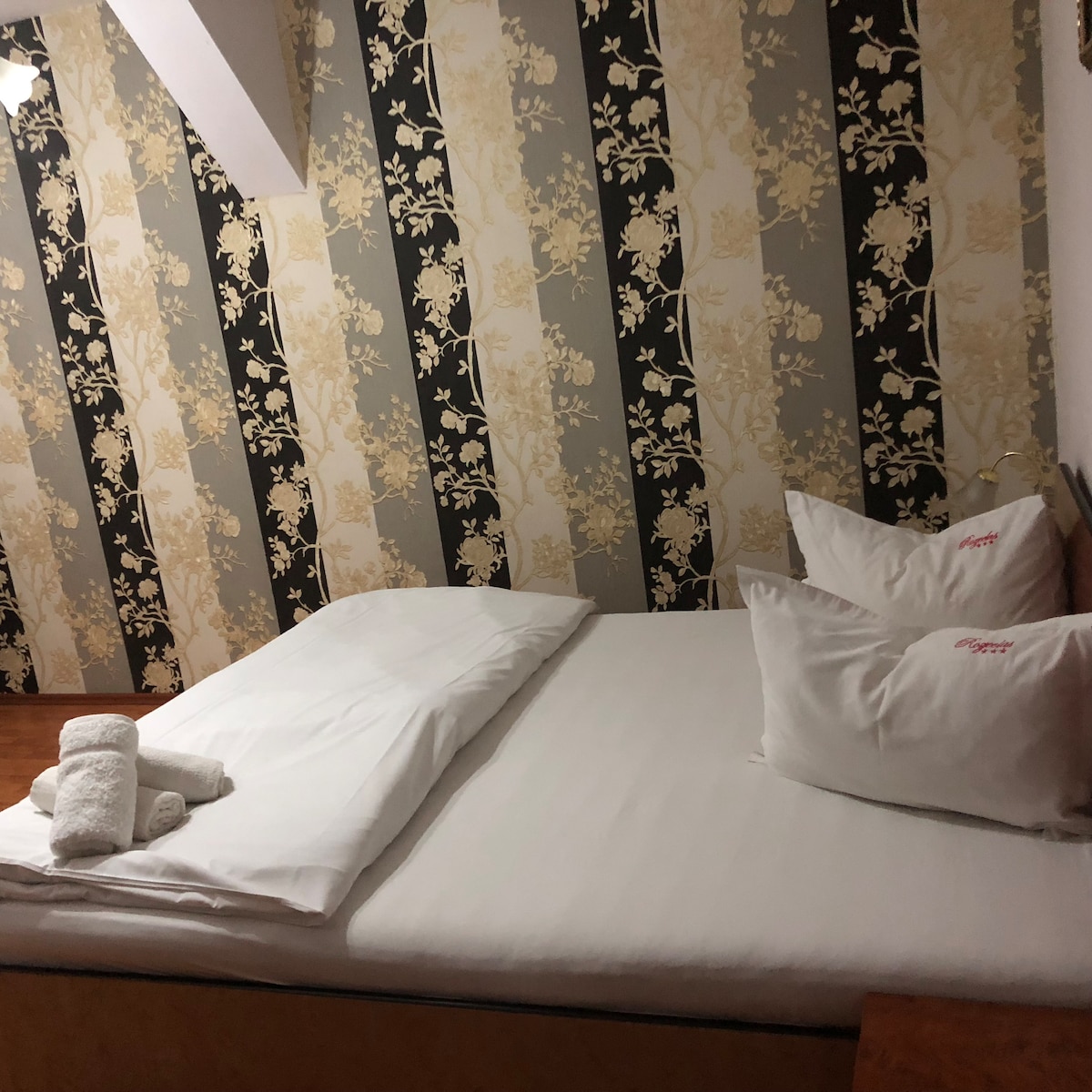 A comfortable sleeping area features a spacious bed with crisp white linens and two decorative pillows. Soft towels are neatly arranged on the bed. The backdrop is adorned with floral wallpaper in shades of gold and black, adding a touch of elegance to the room.