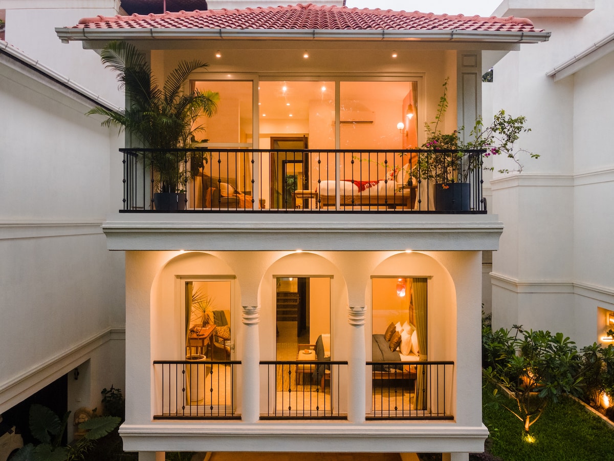 The villa exterior showcases a two-story structure with a terracotta-tiled roof. Balconies are adorned with potted plants, and large glass doors allow visibility into the welcoming interior spaces. Soft lighting highlights the architectural details and surrounding lush greenery.