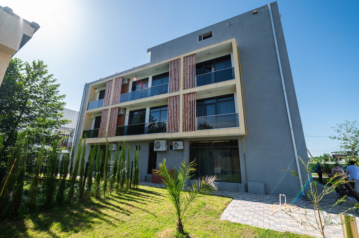 The exterior of the hotel is highlighted by a modern façade with multiple balconies. A well-maintained garden featuring green shrubs and palm trees frames the building. Sunlight illuminates the structure against a clear blue sky, providing a fresh and bright ambiance.